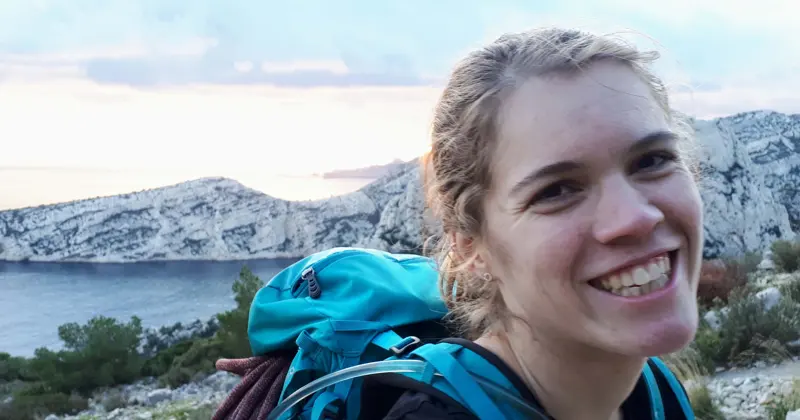 Een glimlachende vrouw met een rugzak staat buiten, met op de achtergrond rotsachtige heuvels, groene bomen en water, terwijl de zon ondergaat.
